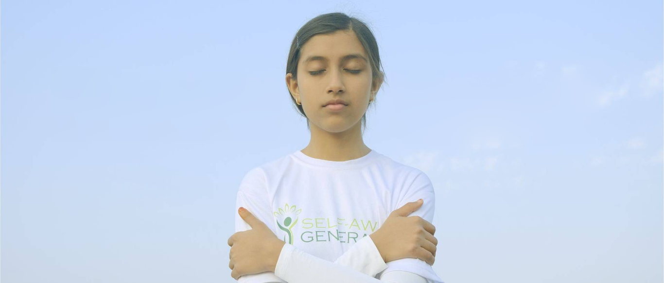 A girl in a white Self-aware Generation shirt stands with her eyes closed and her arms crossed over her chest against a pale blue sky.