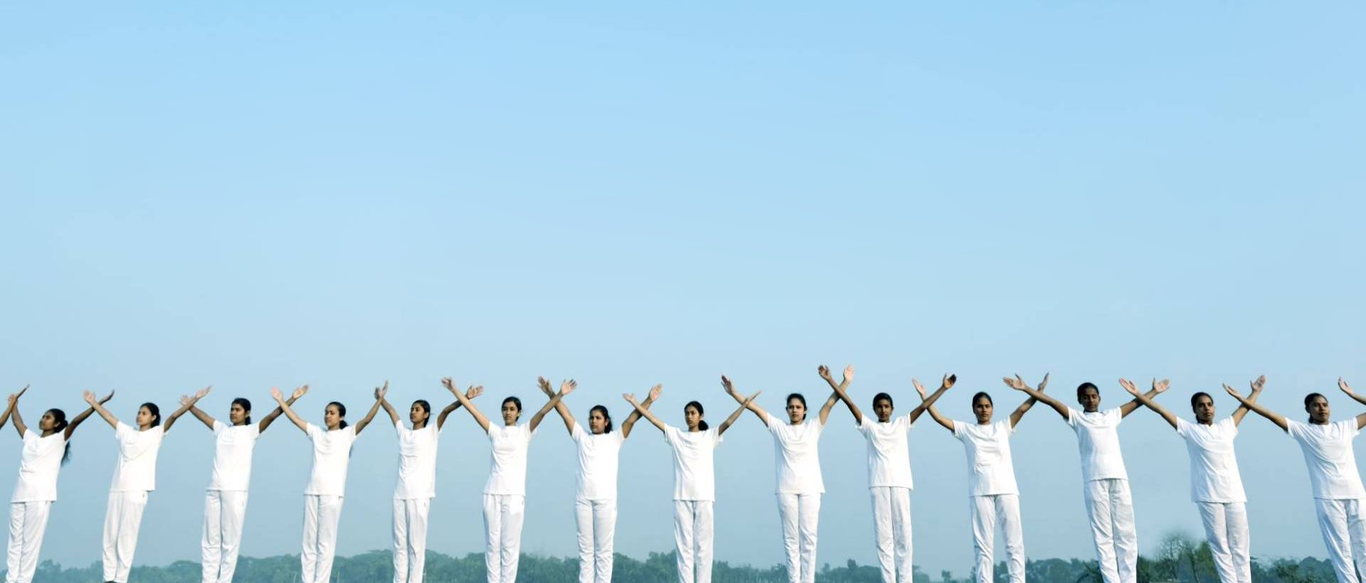 A wide row of young people in white standing across an open field at dawn, arms raised and crossed above their heads.